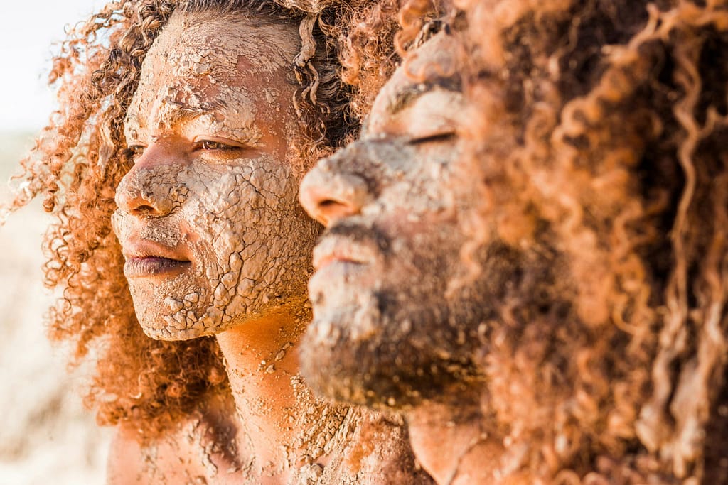 Close-up of two individuals with mud masks and curly hair enjoying daylight outdoors.